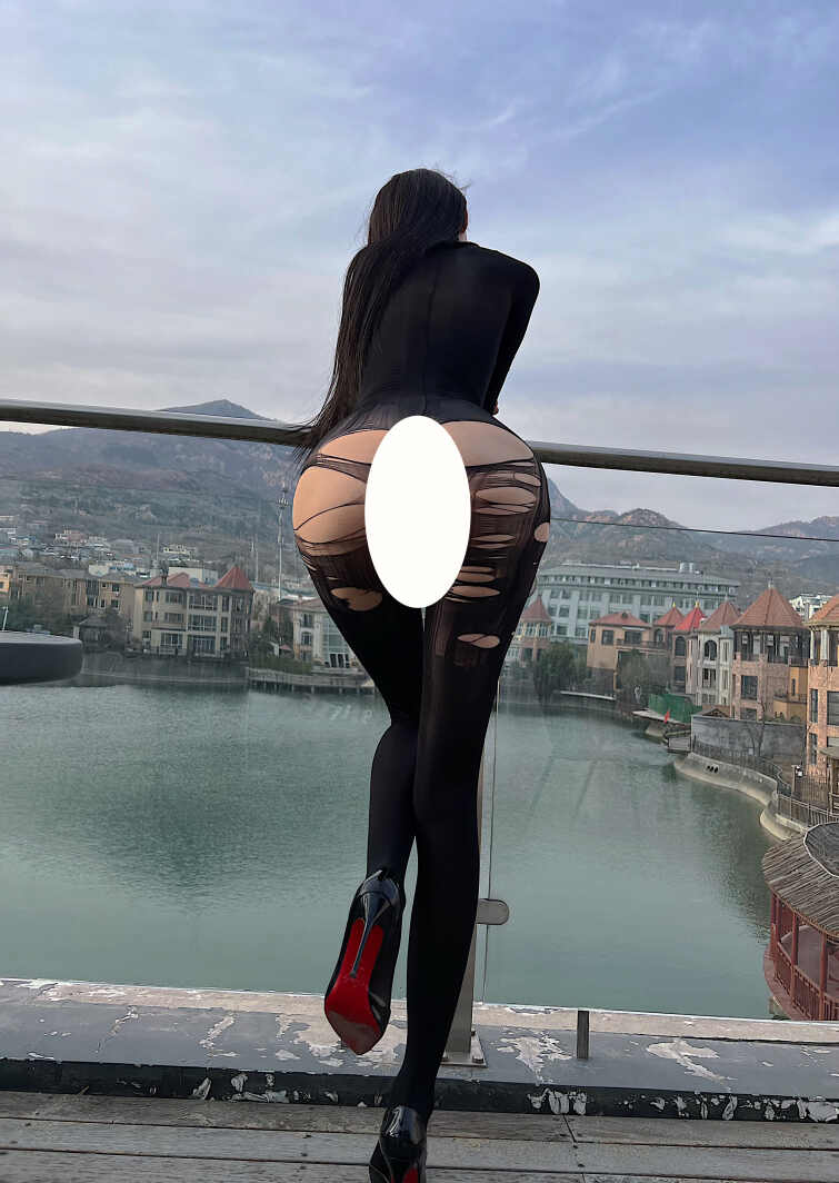 模特脸红Dearie身穿黑色破洞连体衣，背对镜头倚靠天台栏杆，脚穿黑色红底高跟鞋（部分区域已遮挡）