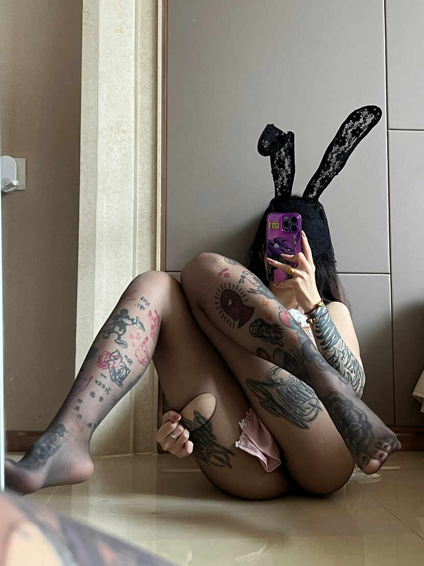 模特卡通百科老王戴黑蕾丝兔耳，穿粉色围裙装和纹身图案丝袜，坐地板镜前自拍，手臂纹身明显