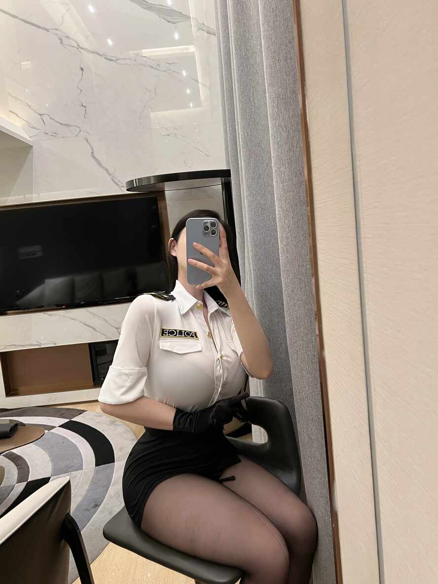 模特左公子666身穿白色制服衬衫、黑色包臀裙和黑色连裤袜，戴黑色手套，坐在椅子上对着镜子自拍
