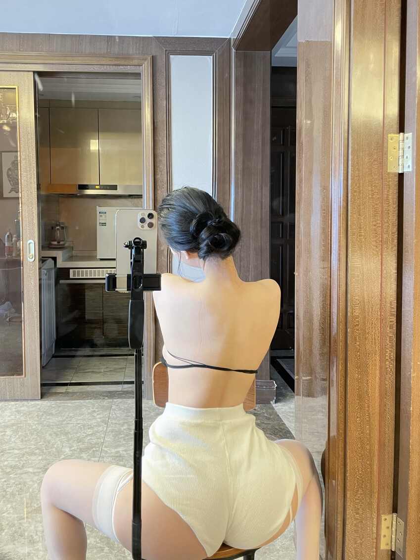 模特左公子666梳丸子头，穿黑色露背上衣/连体衣和白色华夫格短裤，背对镜头坐在椅子上，对着镜子自拍