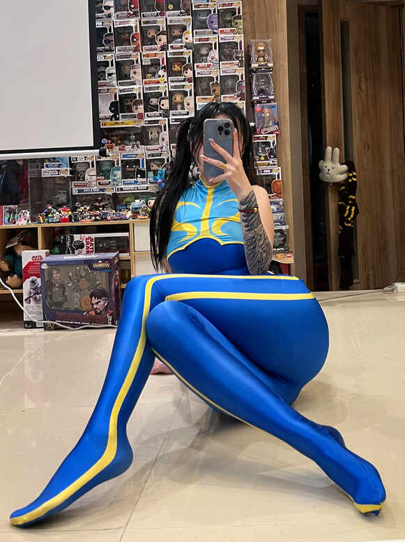 穿着春丽cosplay服装、带有全身纹身的双马尾女子坐在光亮地板上，双腿伸展，手持手机自拍。