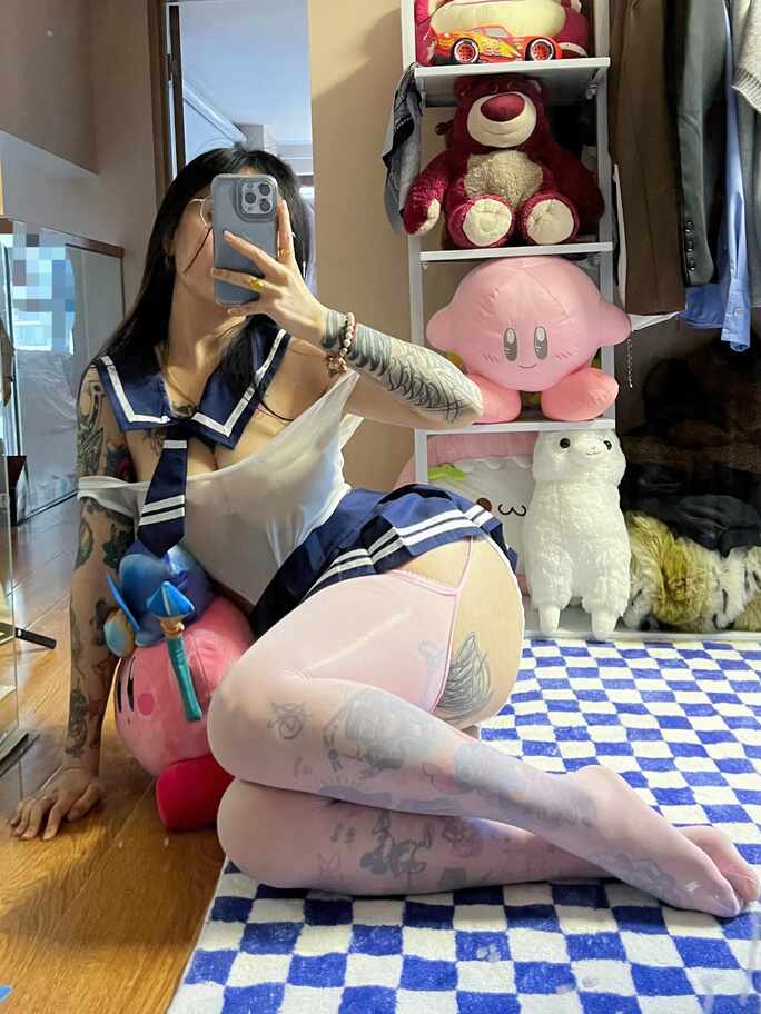 模特卡通百科老王穿水手服上衣、海军蓝短裙和纹身图案丝袜，戴眼镜，侧后方跪姿在镜子前自拍，展示臀部与腿部线条