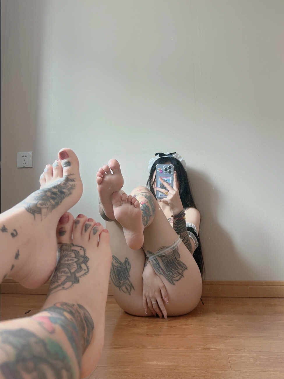 卡通百科老王记录，遍布纹身的女子身穿黑色女仆风比基尼，头戴女仆发箍，仰卧在木地板上，双脚赤裸举向空中，对镜自拍，手机遮脸，展现纹身。