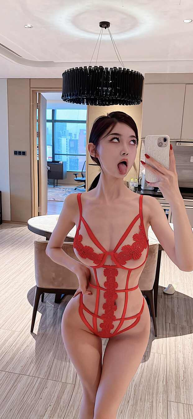 脸红Dearie身穿红色蕾丝刺绣镂空连体衣，在室内对镜自拍，手机遮挡部分脸部，做出翻白眼伸舌头的俏皮表情。