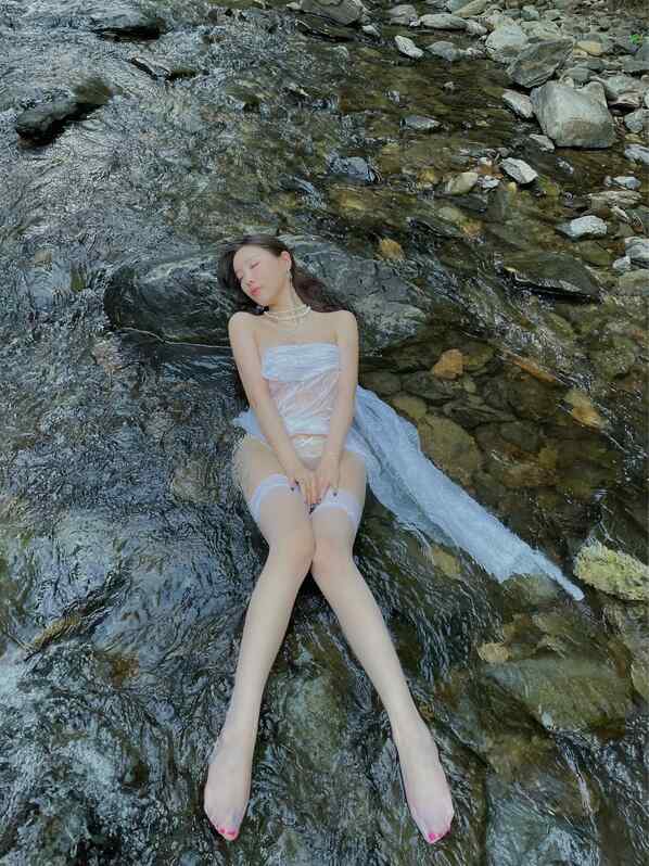 杨晨晨身着飘逸白纱裙，如水精灵般躺卧于清澈溪流的岩石上，展现“上水间”主题的空灵唯美。