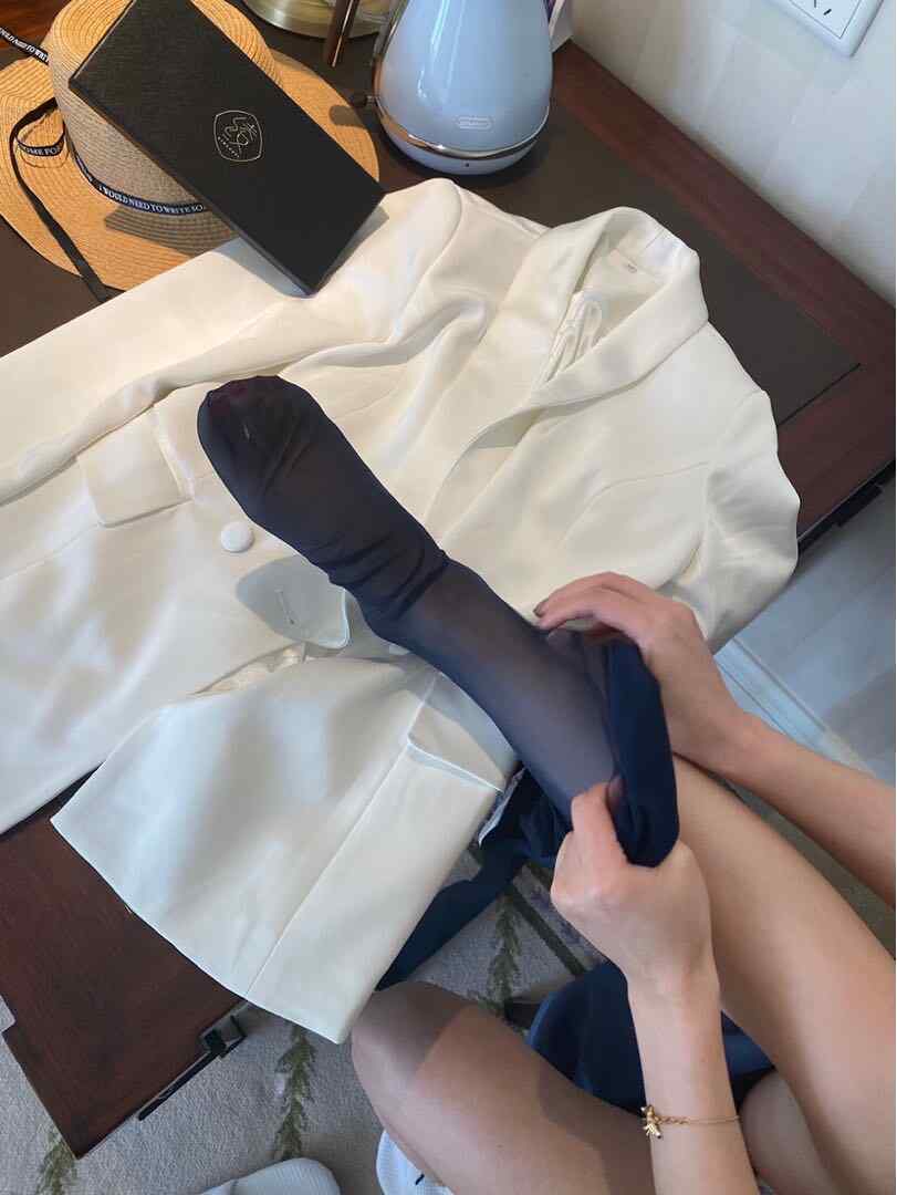 July芝岚正将一只深蓝色/黑色透明丝袜穿过脚踝，旁边放着一件白色西装外套和一顶草帽。
