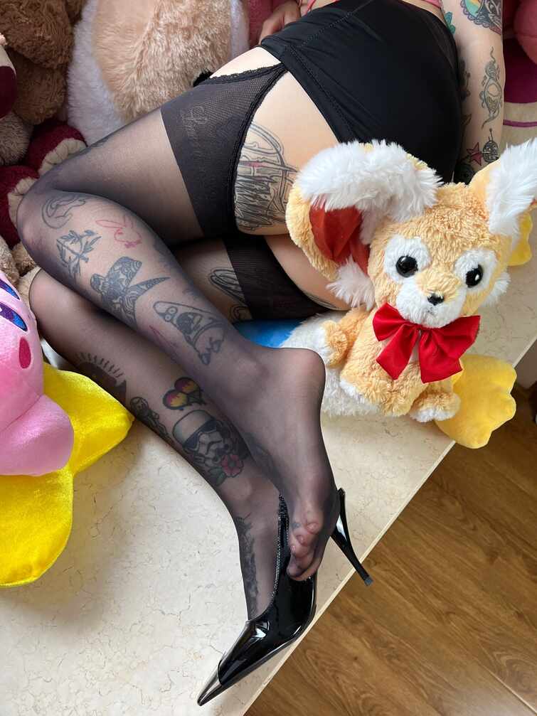 卡通百科老王镜头下，遍布纹身的女子腿部穿着黑色印花长筒袜和黑色高跟鞋，与可爱的狐狸毛绒玩偶一同出现在画面中。